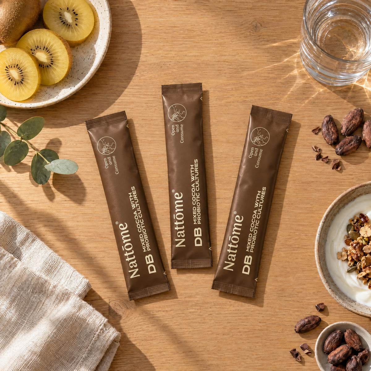 Three Nattōme DB Cocoa sachets on a breakfast table with golden kiwifruit, yoghurt and a glass of water